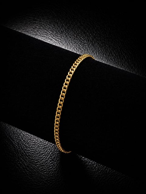 Gold curb chain bracelet displayed on black textured surface with dramatic lighting