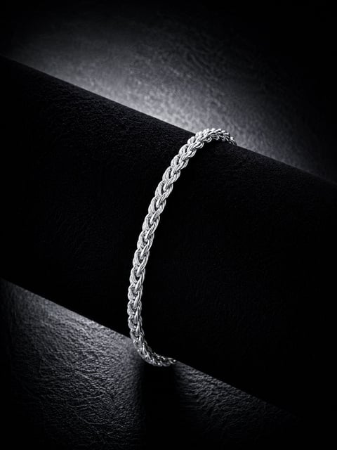 Silver braided chain bracelet displayed on black geometric surfaces with dramatic lighting