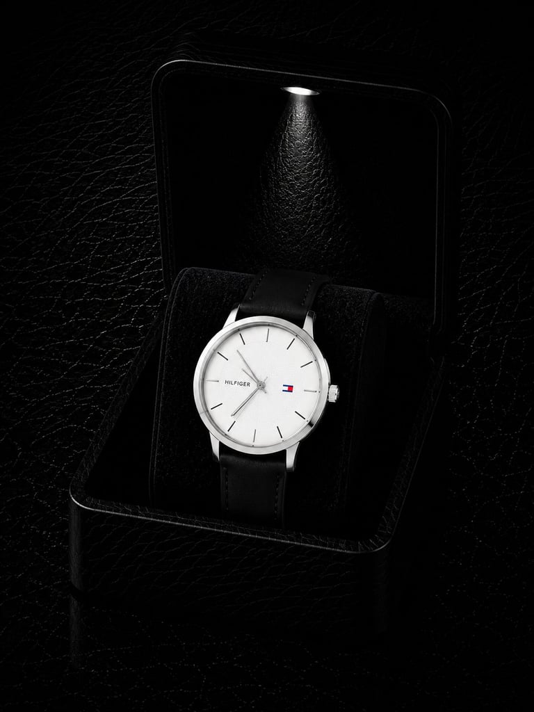 Silver and white Tommy Hilfiger watch with black leather strap displayed in an open black box with spotlight lighting